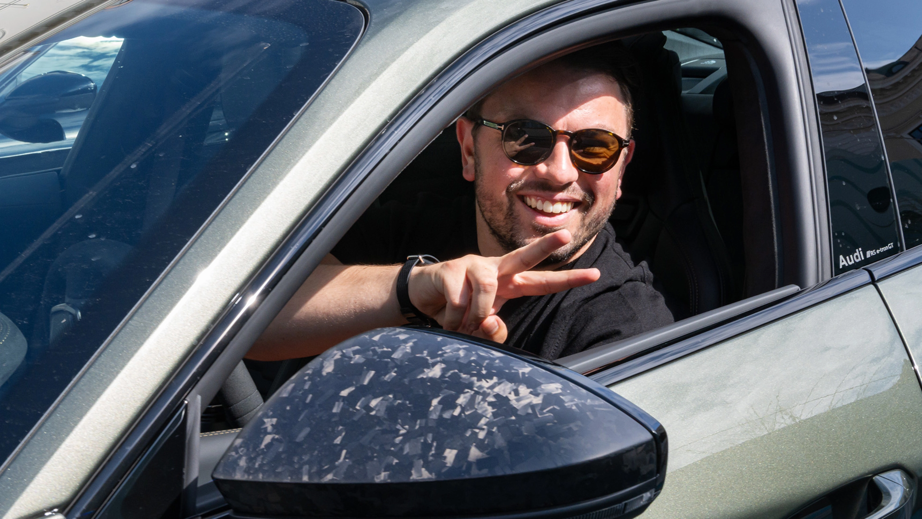 Mann mit Sonnenbrille lächelt und gestikuliert vom Fahrersitz eines Autos.