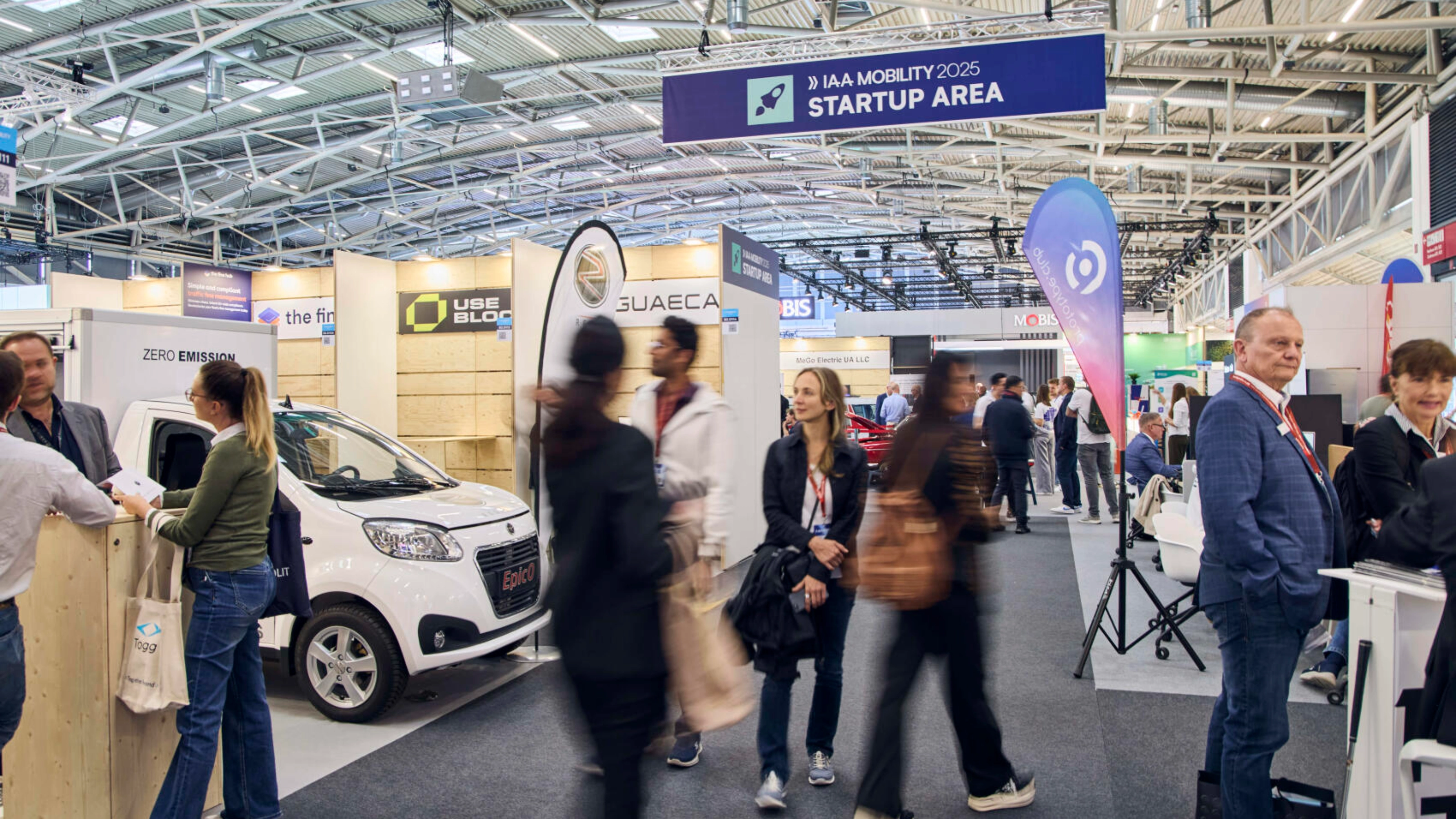 Menschen gehen durch die IAA MOBILITY 2025 Startup Area mit Ständen und E-Fahrzeugen.