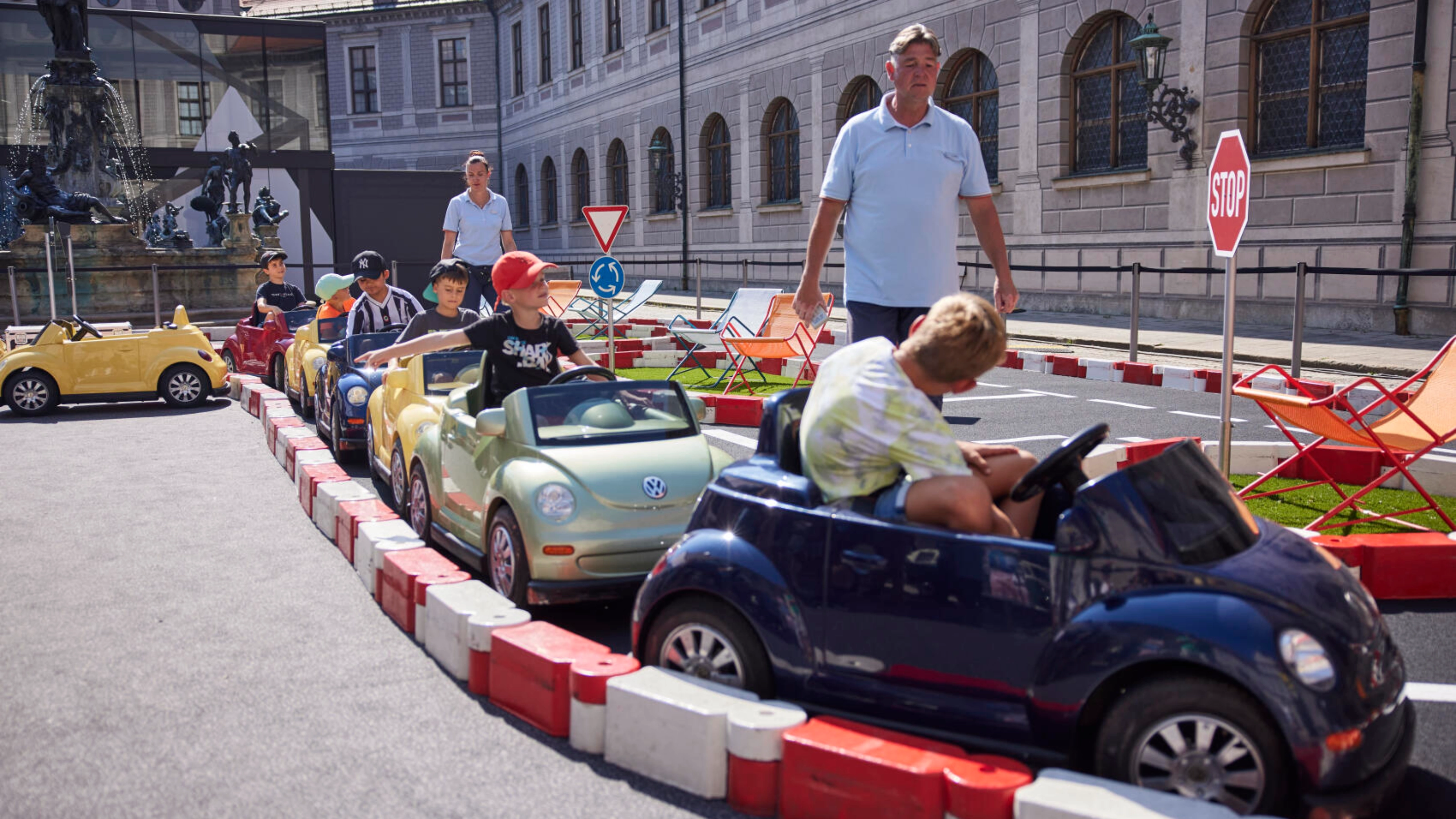 Children drive small electric toy cars on a mini road setup at an outdoor event, while adults and other kids watch from the side.