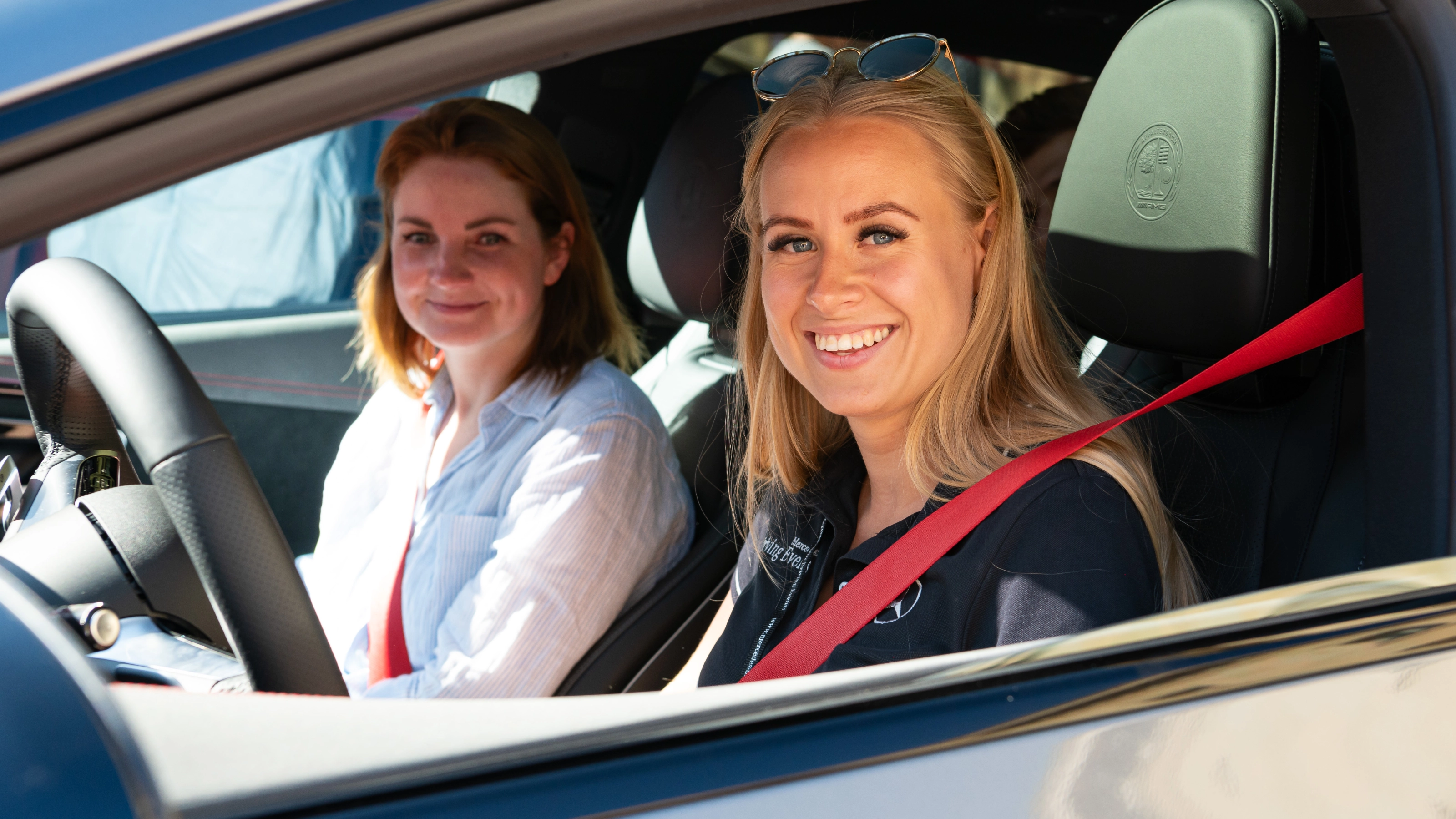 Two smiling women sit in the front seats of a car, both wearing seat belts.