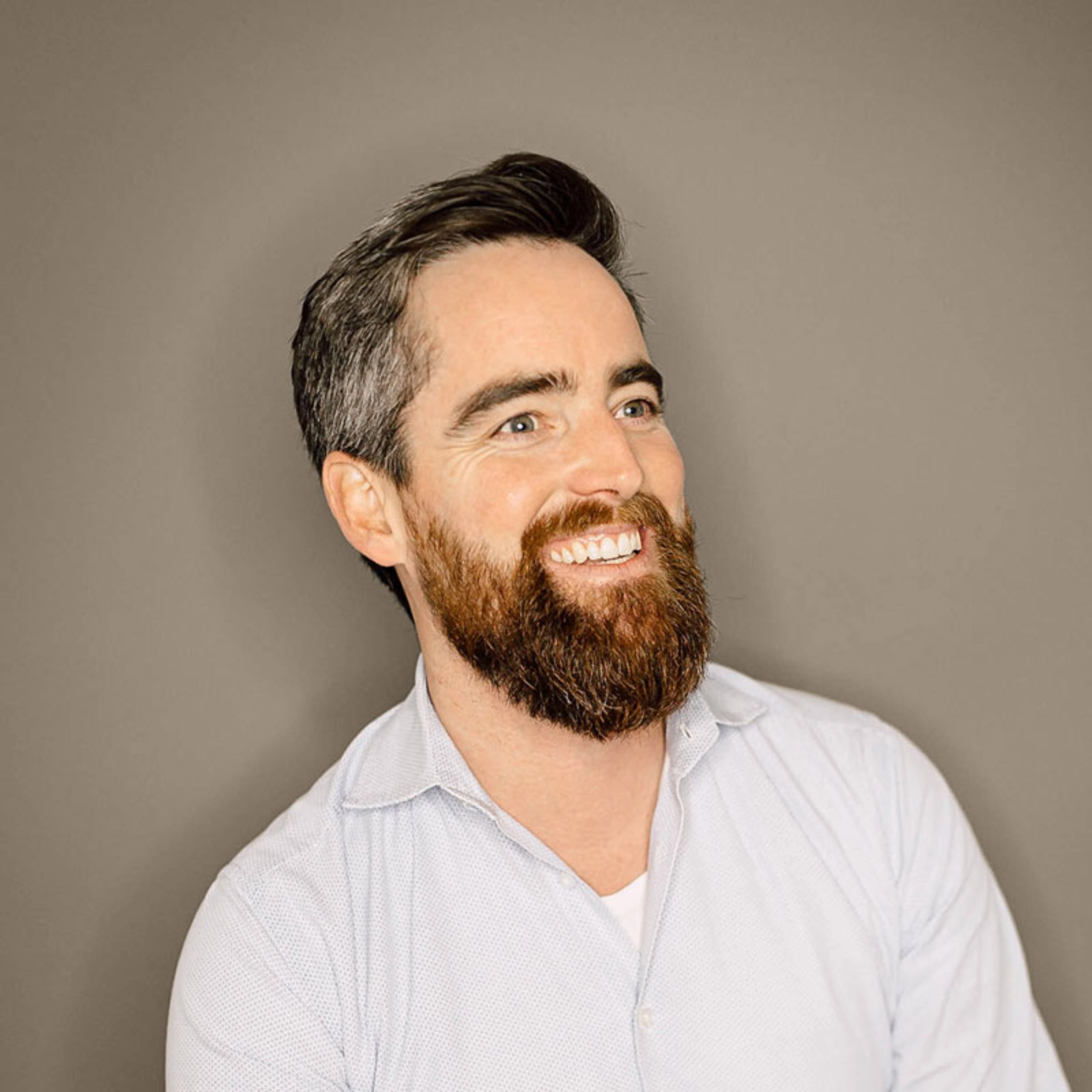 Smiling man with beard in light shirt against neutral background.
