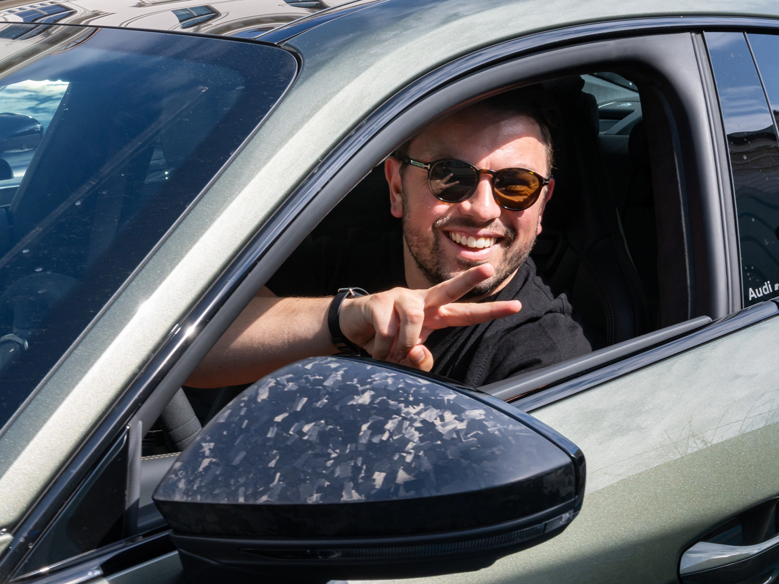 Mann mit Sonnenbrille lächelt und gestikuliert vom Fahrersitz eines Autos.