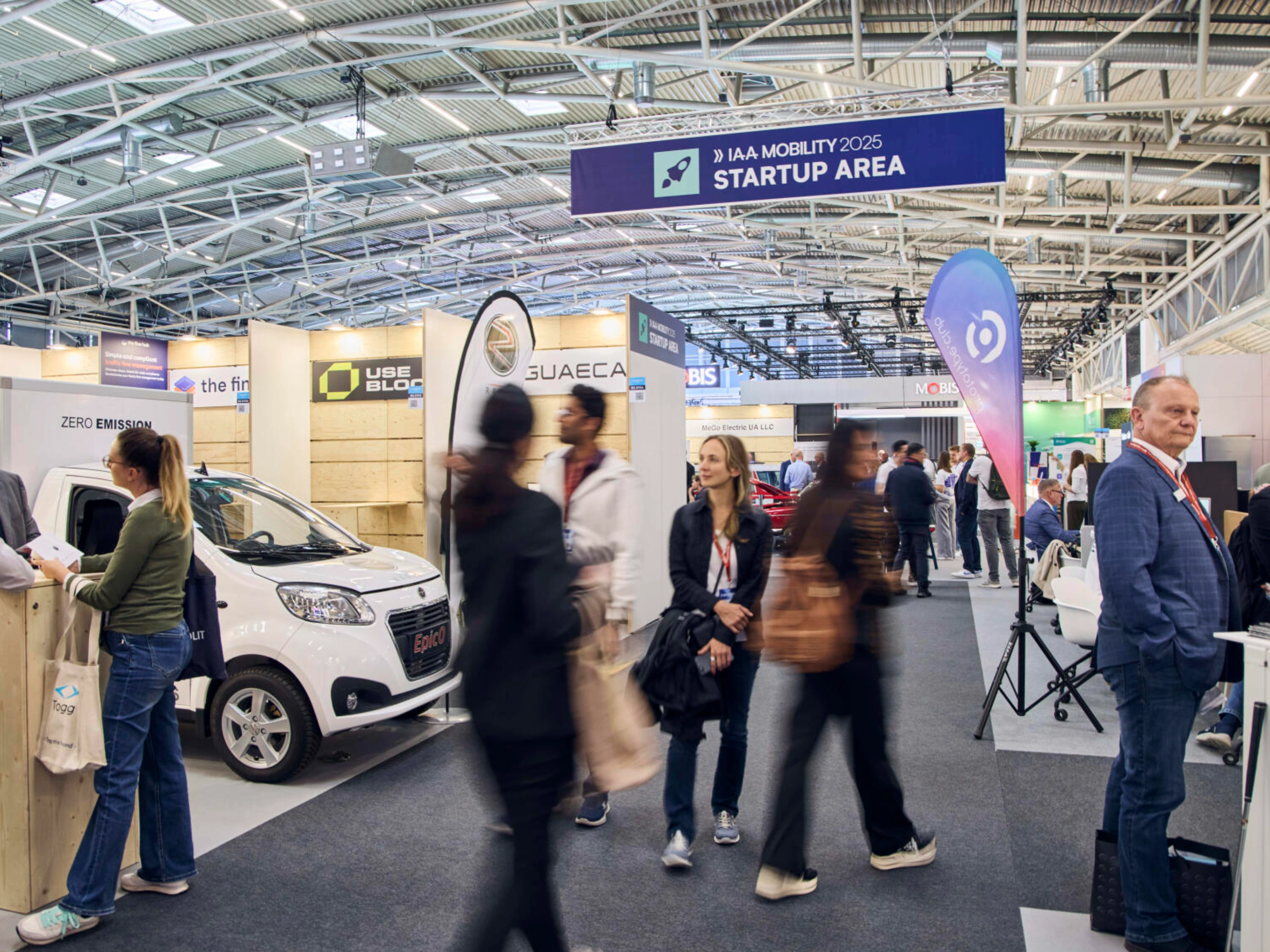 Menschen gehen durch die IAA MOBILITY 2025 Startup Area mit Ständen und E-Fahrzeugen.