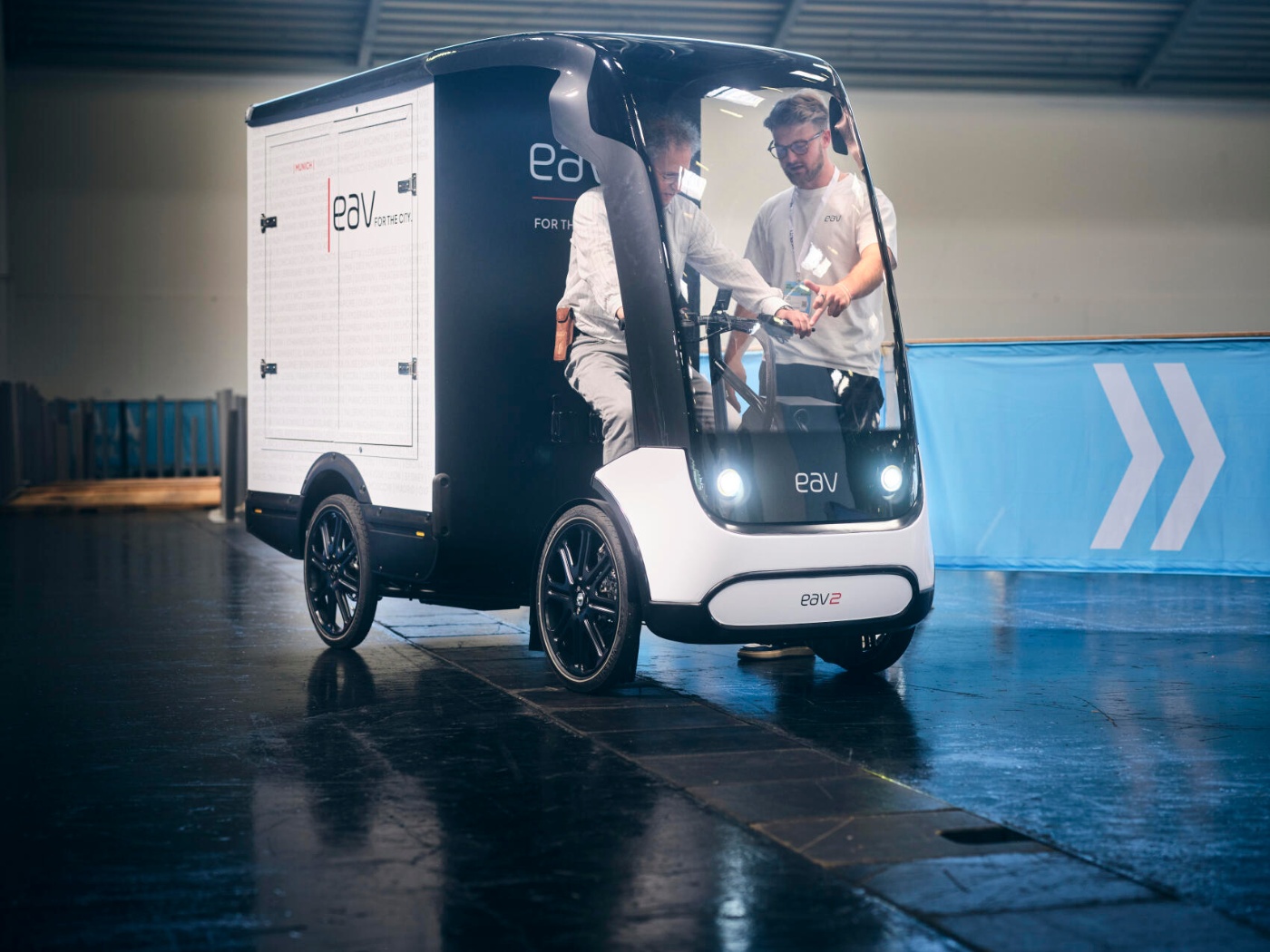 Zwei Männer testen ein kompaktes Elektro-Lieferfahrzeug mit der Aufschrift „eav“ in der Halle.