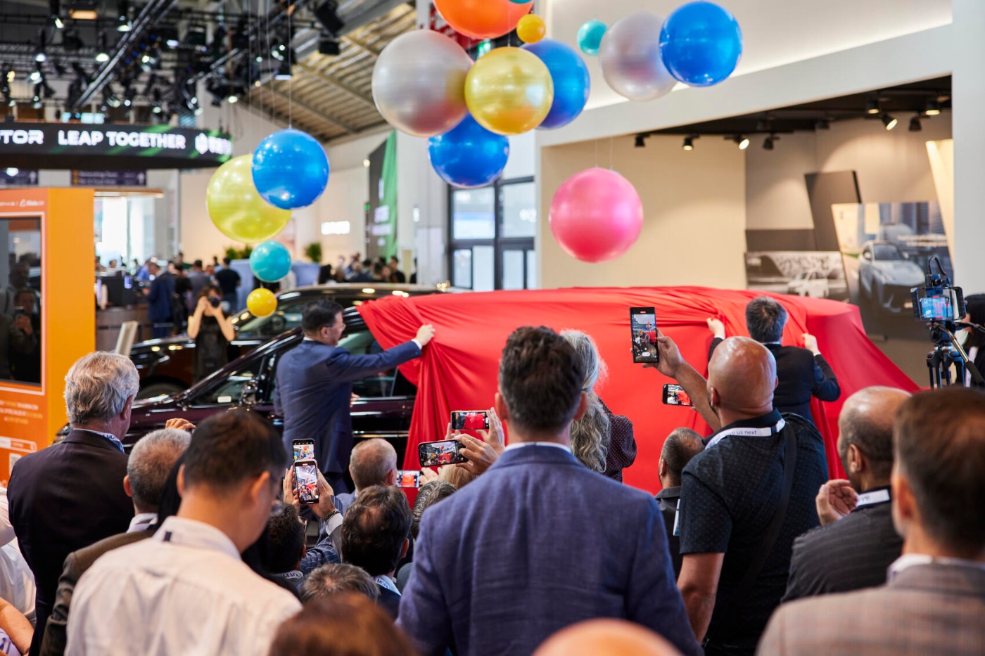 A crowd watches and films the unveiling of a car covered with a red cloth at an event.