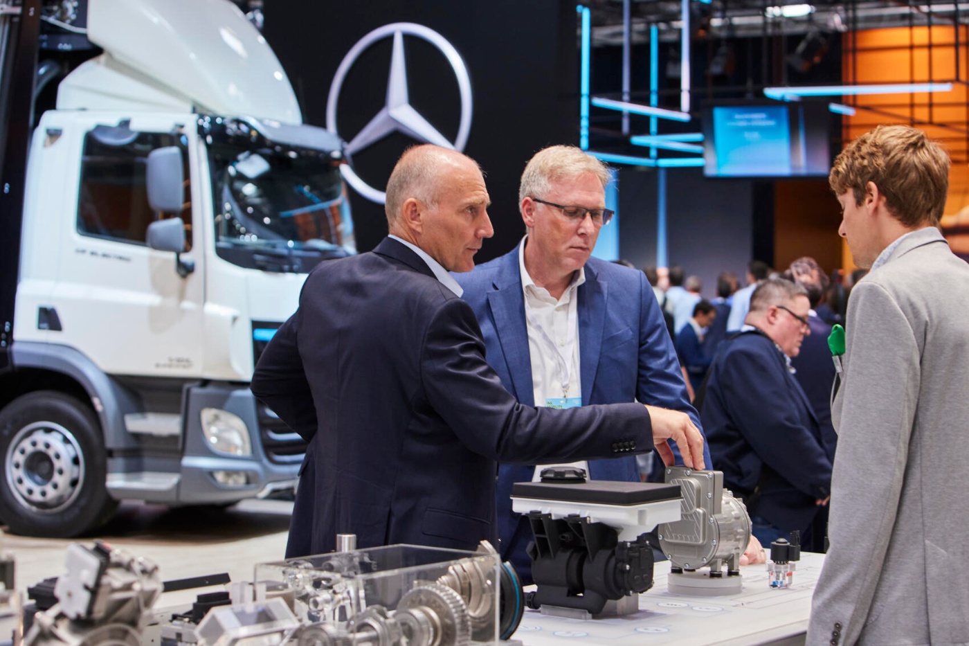 Three men discuss automotive components at a trade fair booth in front of a Mercedes-Benz truck.