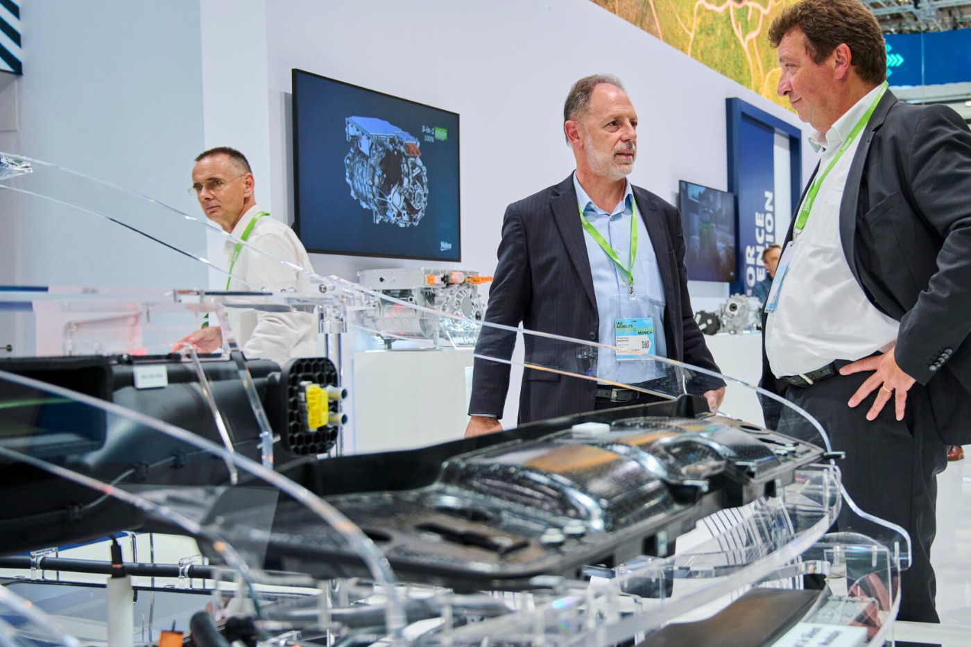 Three men discuss automotive components at a trade fair booth with technical displays.