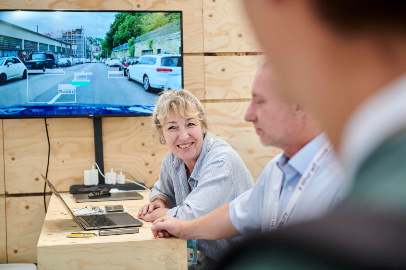 A woman smiles during a tech demo with a laptop and screen showing a street scene analysis.