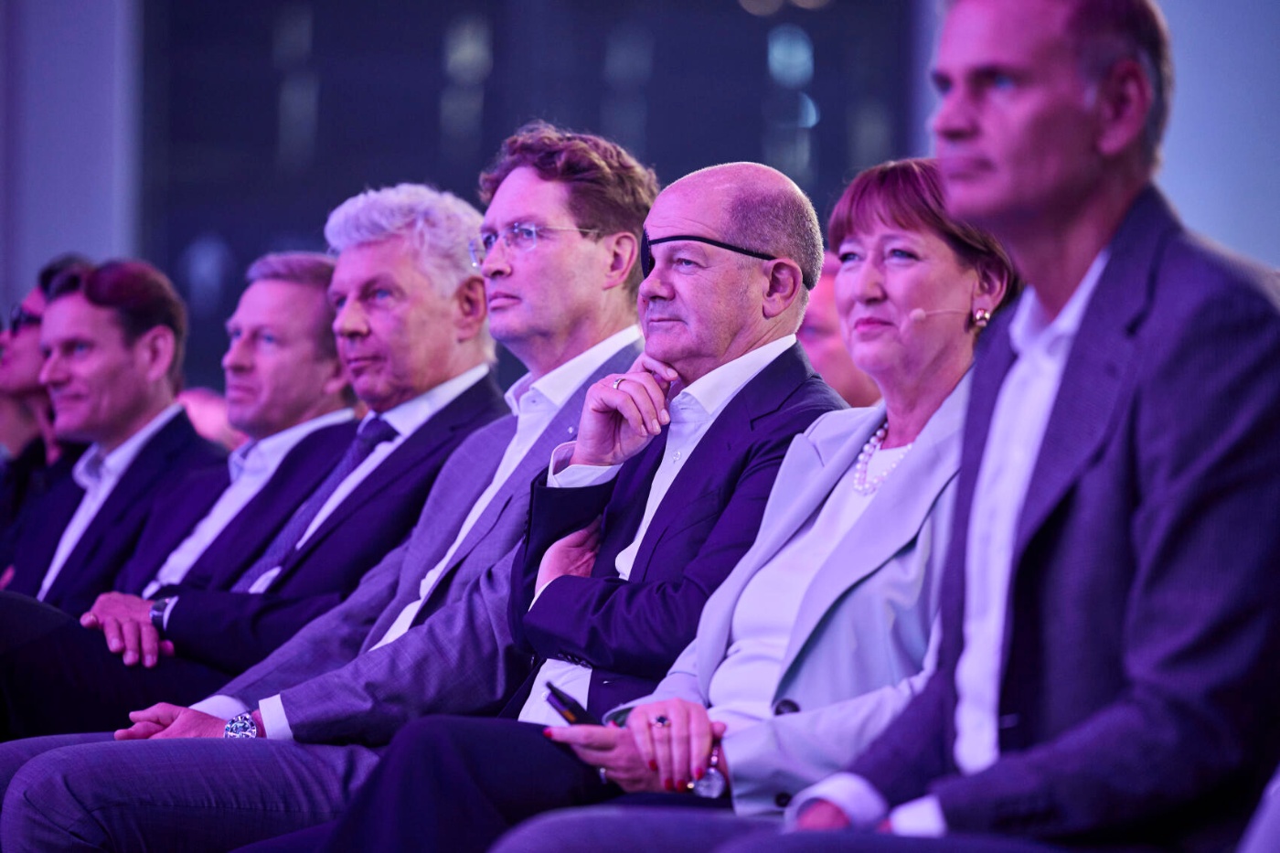 A row of formally dressed people sits attentively at a conference under purple lighting.