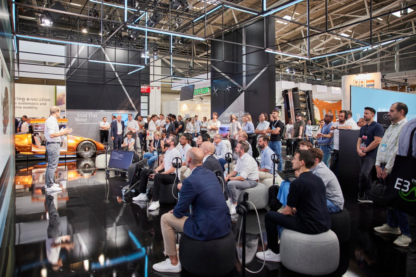 Audience listens to a speaker at a trade fair booth with a futuristic car and large screen display.
