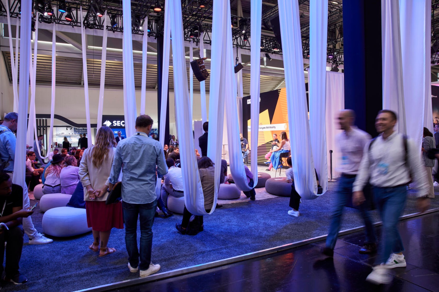 People sit and listen to a panel discussion at a trade fair lounge with hanging fabric decorations.