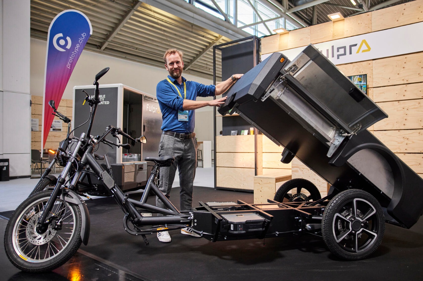 Mann zeigt ein elektrisches Lastendreirad mit kippbarem Container auf einem Messestand.