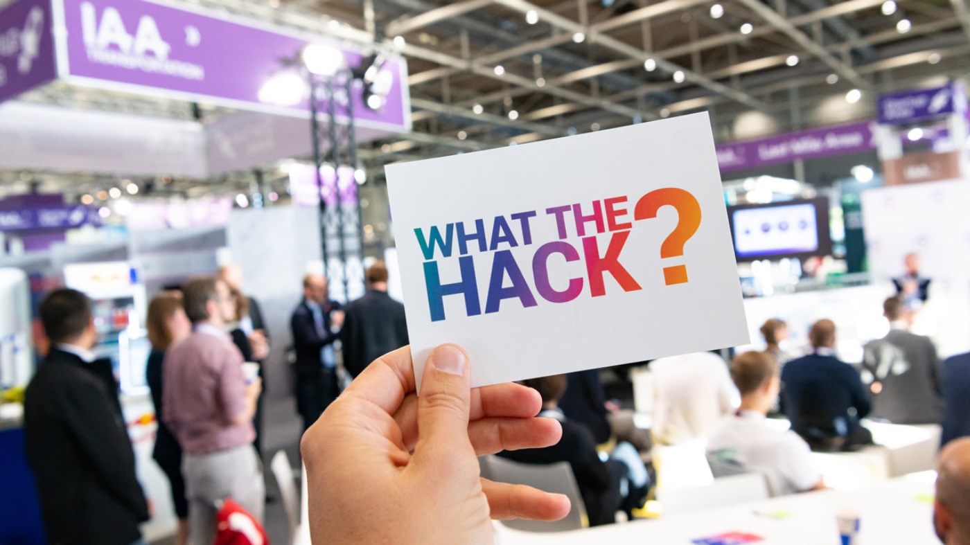 Person holding a card reading "What the Hack?" at a tech event.
