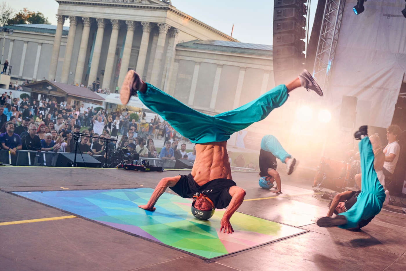 Breakdancers performing headspins and acrobatic moves on an outdoor stage at IAA Mobility 2023, with a large audience and historic building in the background.