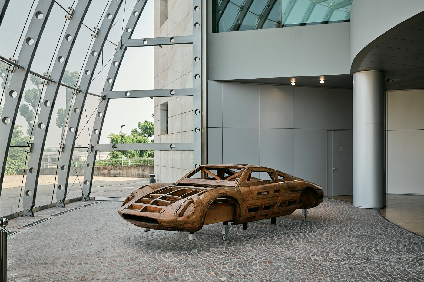 Wooden car body model displayed in a modern glass-walled building.
