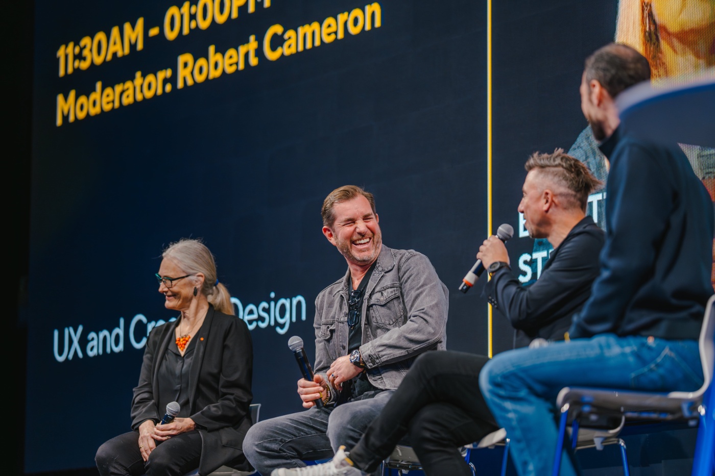 Three people with microphones laugh during a panel discussion on stage.
