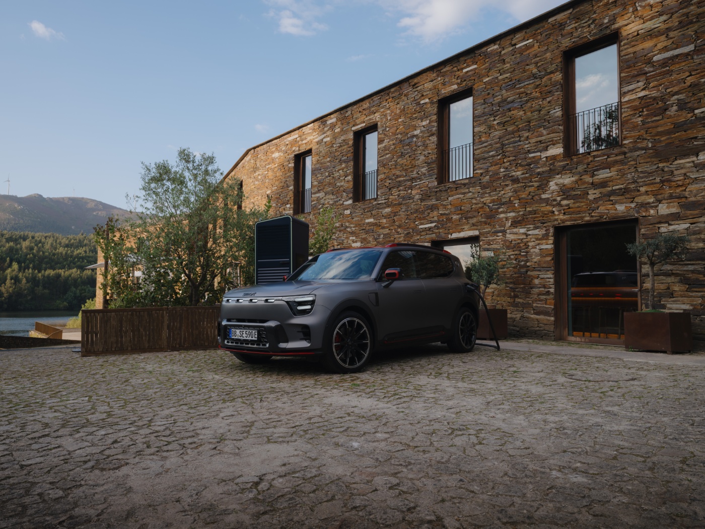 Electric SUV is charging in front of a stone house in a rural, hilly landscape.