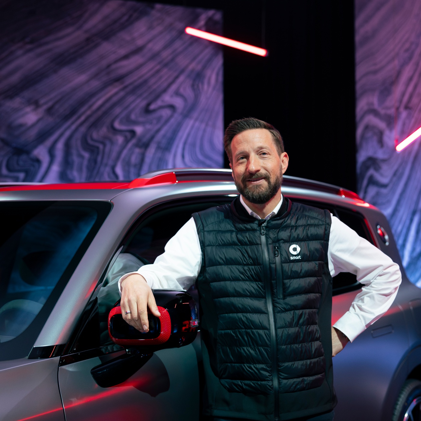 Man in smart-branded vest stands smiling next to an SUV at an indoor event.