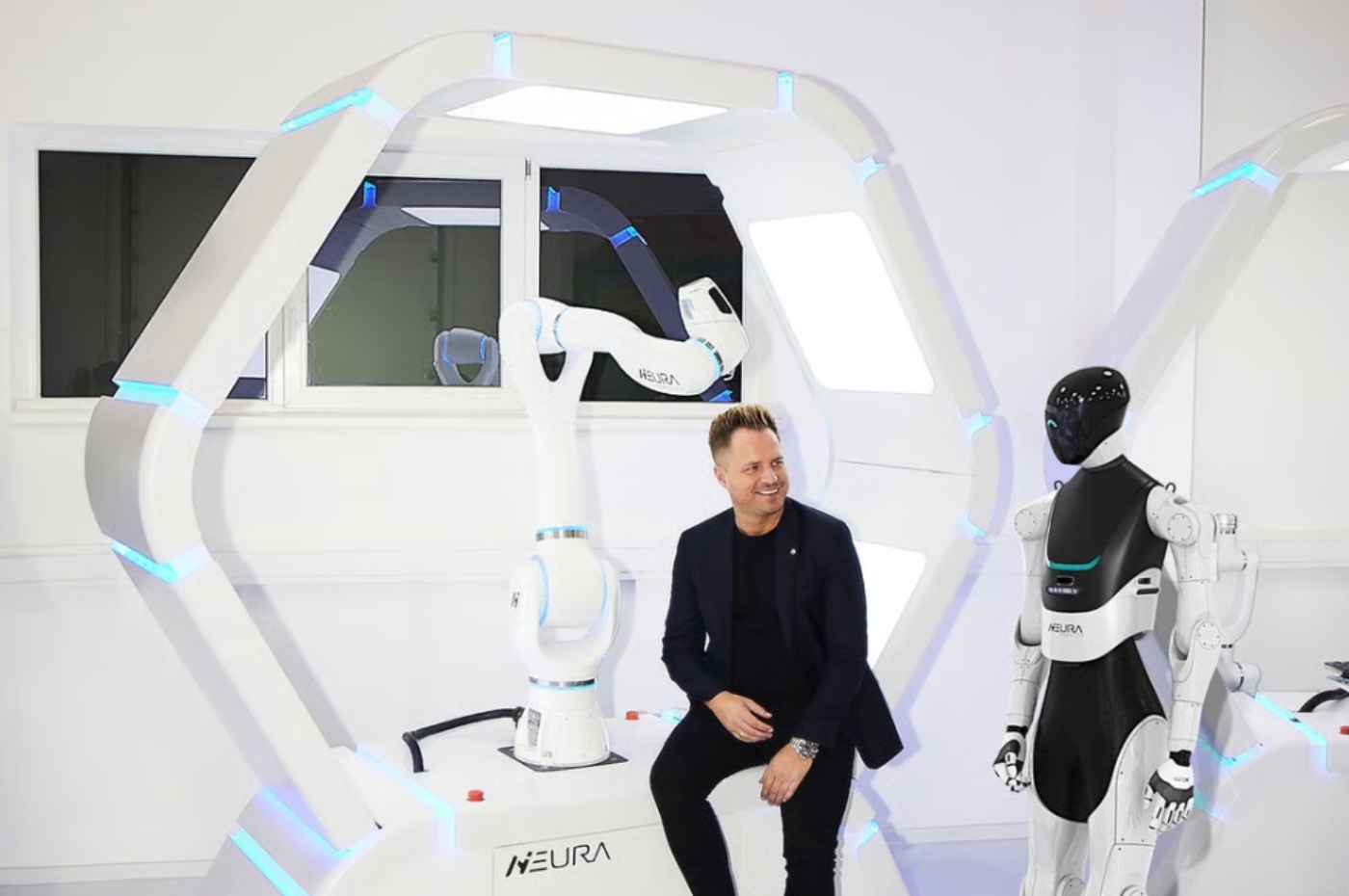 Ein Mann sitzt lächelnd zwischen einem humanoiden Roboter und einem Roboterarm in einem Technikraum.