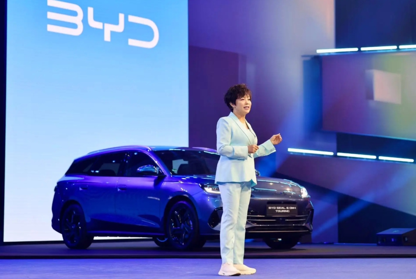 Frau präsentiert auf einer Bühne vor einem BYD Seal 6 DM-i Touring bei blauem Licht.