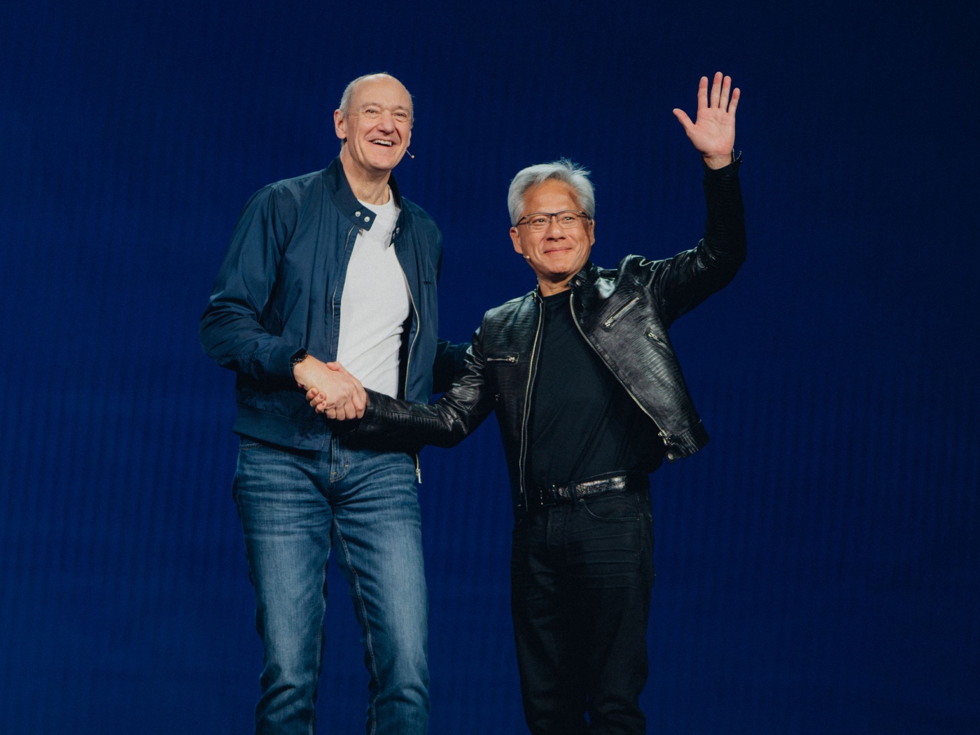 Two men on stage shaking hands, one waving, both smiling in front of a plain blue background.