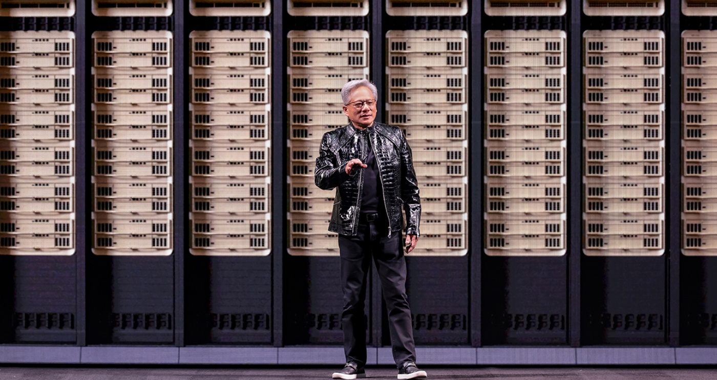 Man stands on stage in front of a large wall of computer servers during a tech presentation.