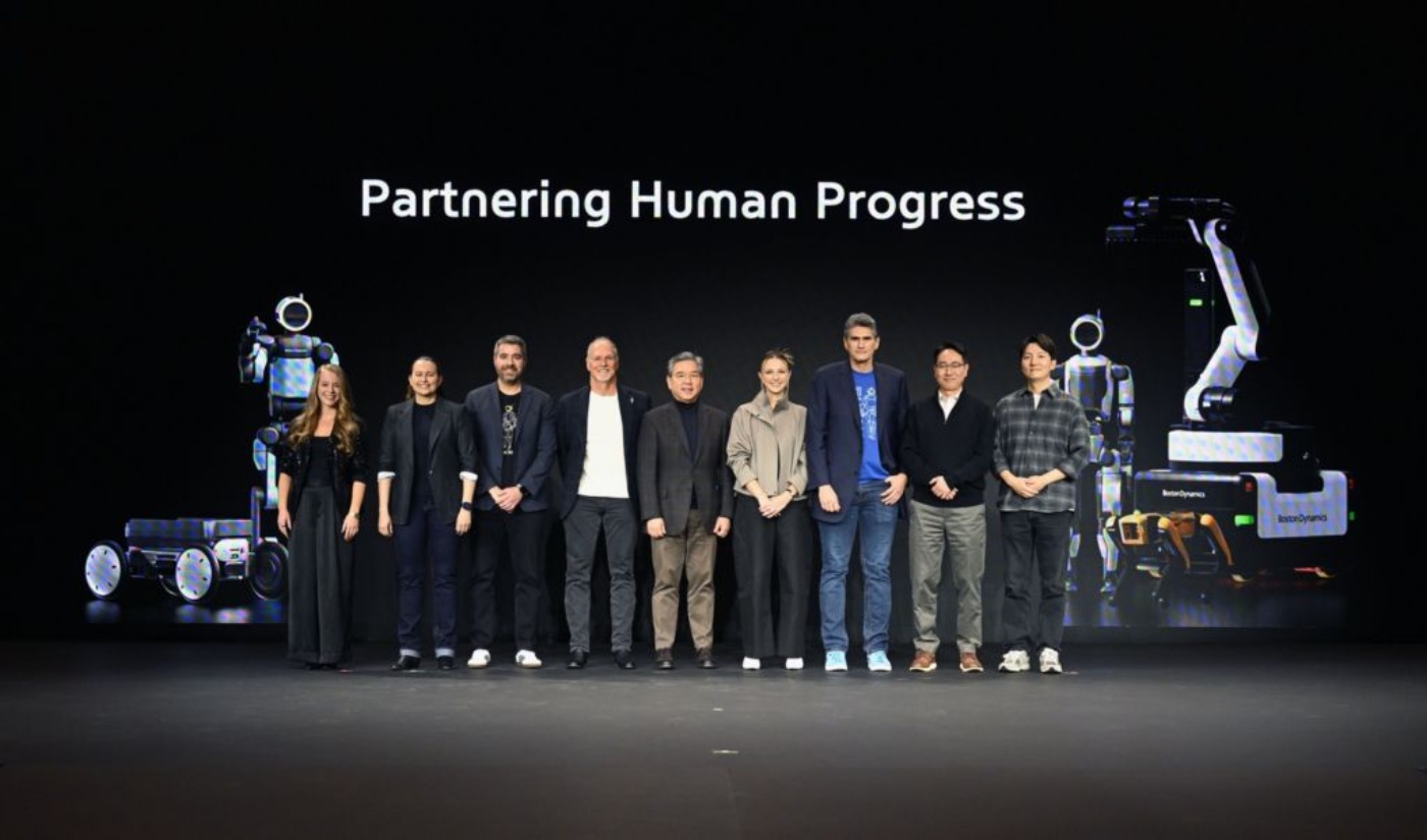 Group of people on stage with robots and the slogan "Partnering Human Progress" in the background.