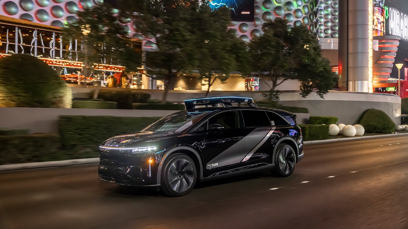 Self-driving electric SUV drives past brightly lit buildings at night in an urban setting.