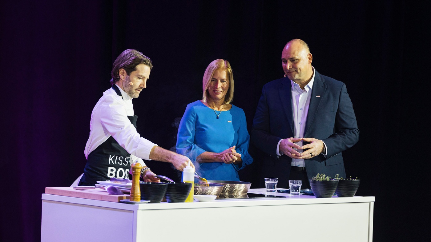 A chef cooks live on stage while two people in business attire watch and smile.