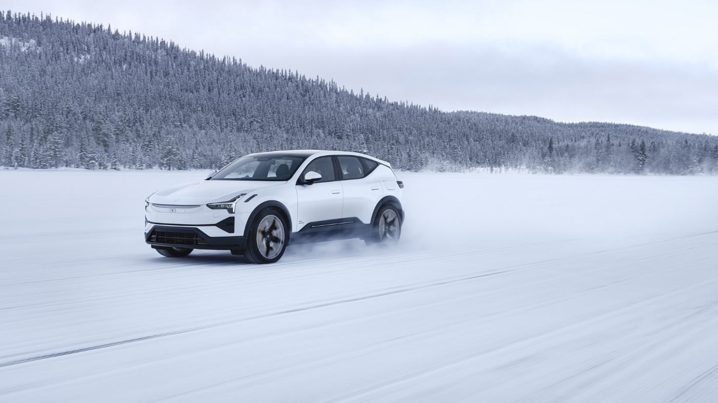 A modern white SUV driving across a snowy landscape, leaving tracks and snow spray behind, with a forested hillside in the background.
