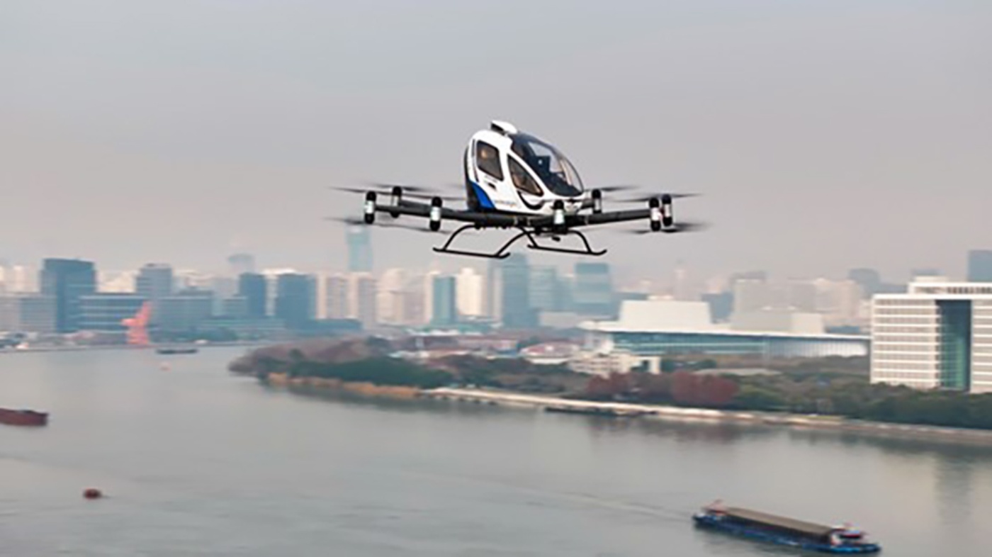 An autonomous passenger drone flying over a river with a modern city skyline in the background.