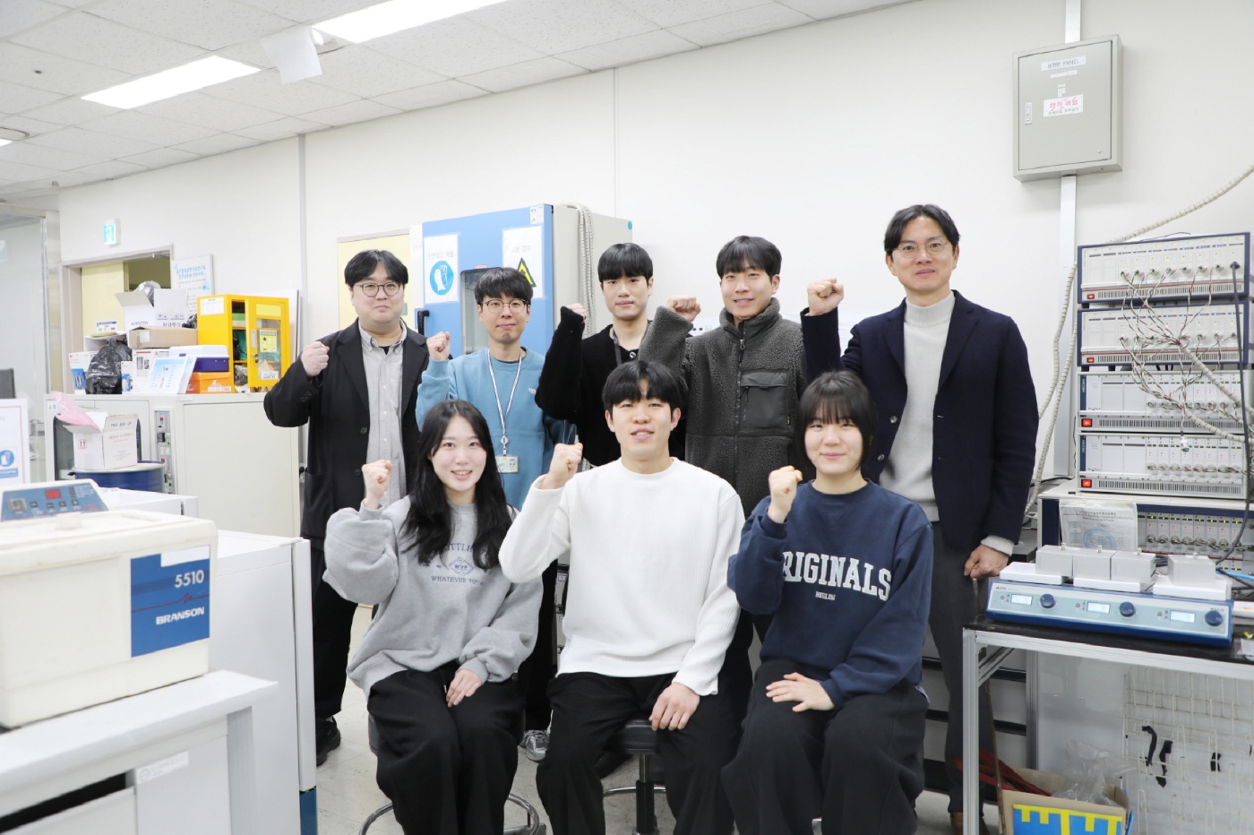 Group of eight young adults posing and smiling in a laboratory, all raising one fist in a gesture of motivation or solidarity.