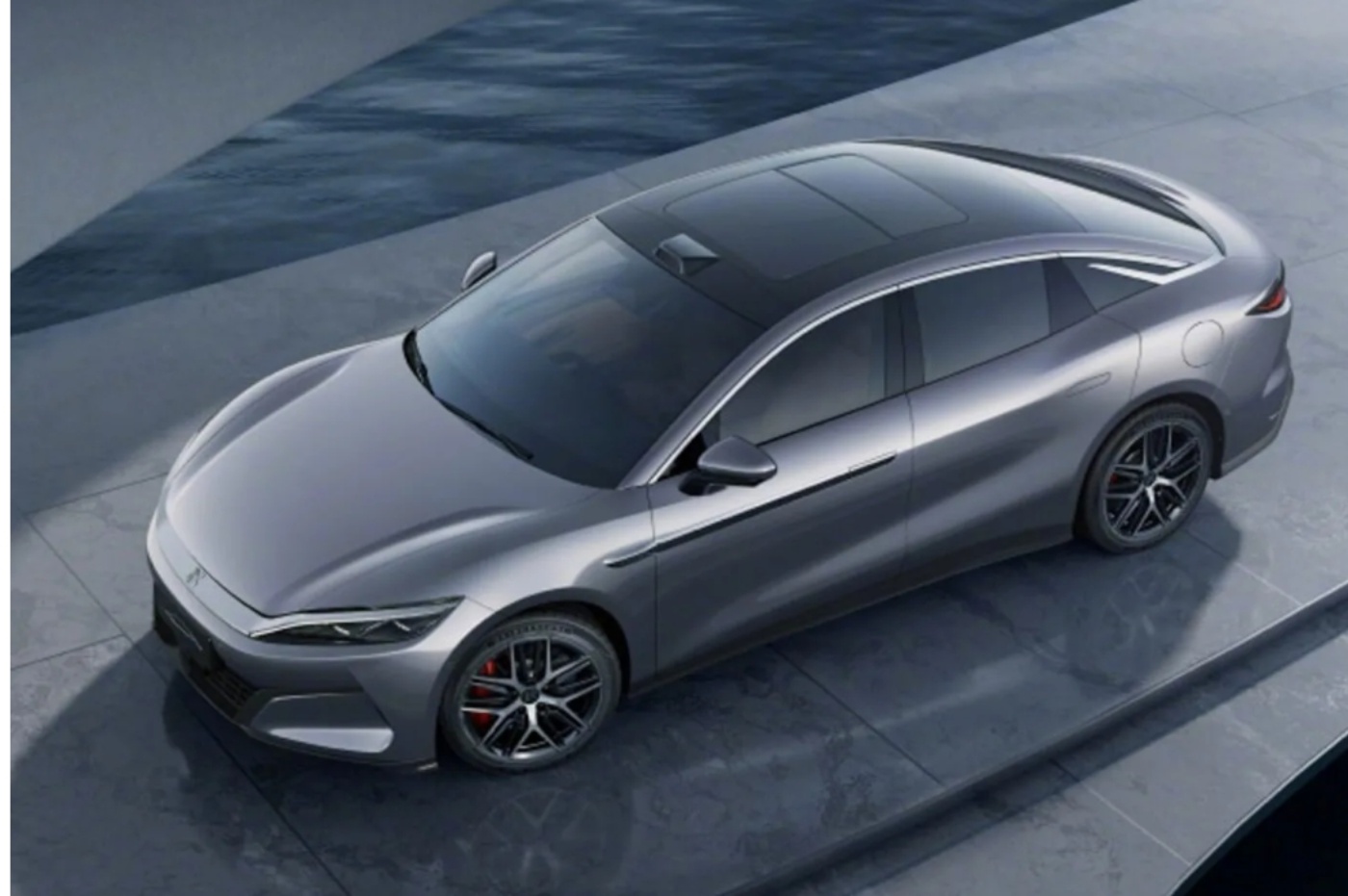 Top view of a modern silver electric sedan parked on a sleek concrete platform near water.