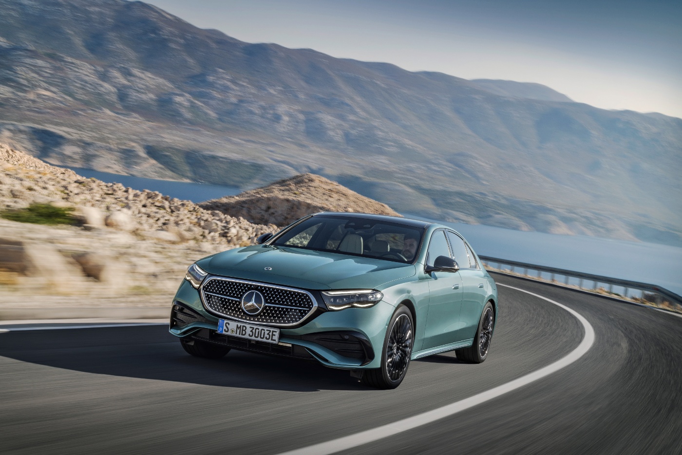 A luxury sedan driving on a winding mountain road with a scenic view of hills and water in the background.