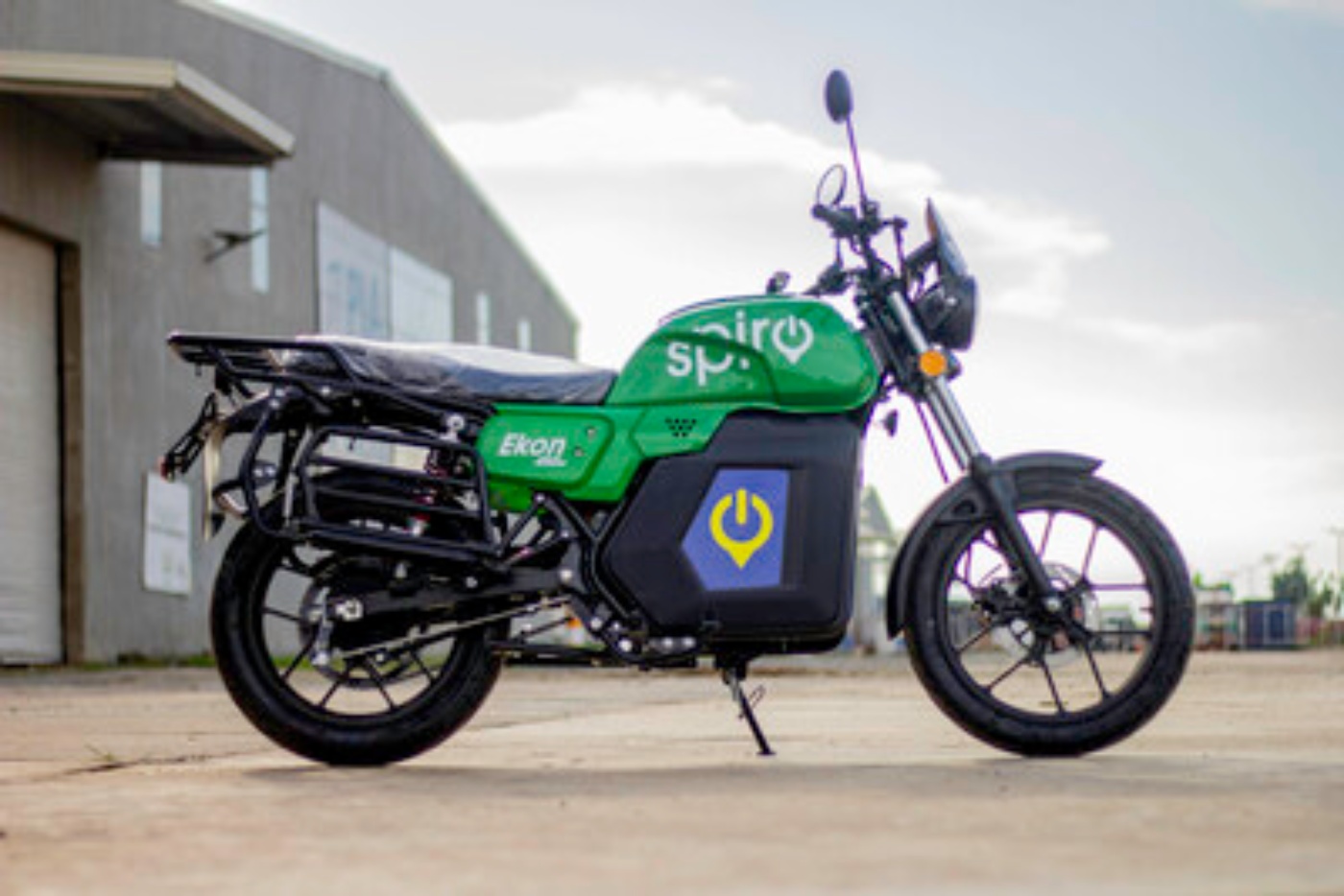 Side view of a green electric motorcycle parked outdoors on concrete, with branding and a power symbol on the battery compartment.