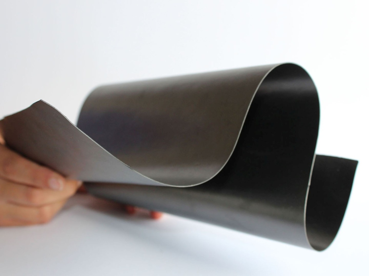 A hand holding a flexible, curved sheet of dark material, possibly a thin electronic or composite layer, against a light background.
