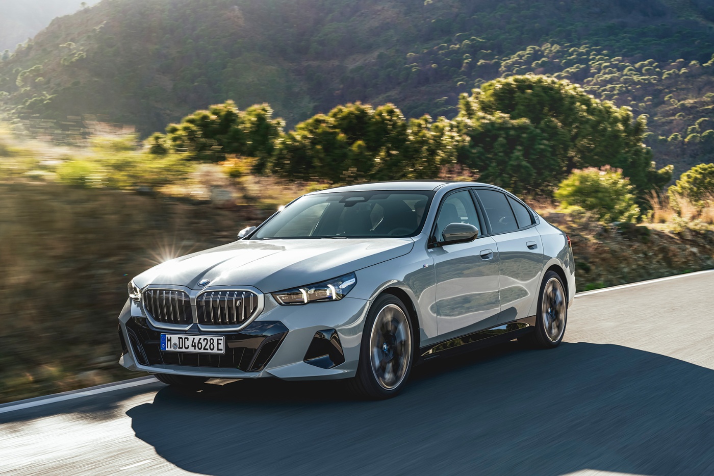 A silver luxury sedan driving on a curving road through a sunlit mountain landscape with trees and hills in the background.