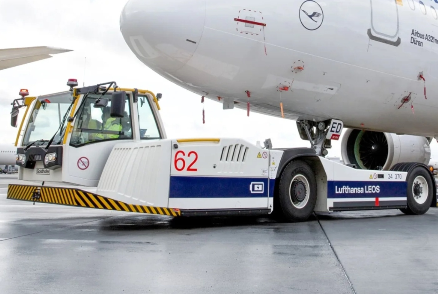 An aircraft tug marked "Lufthansa LEOS" and "Goldhofer" is positioned under the nose gear of an Airbus A321, preparing for pushback on a wet runway.