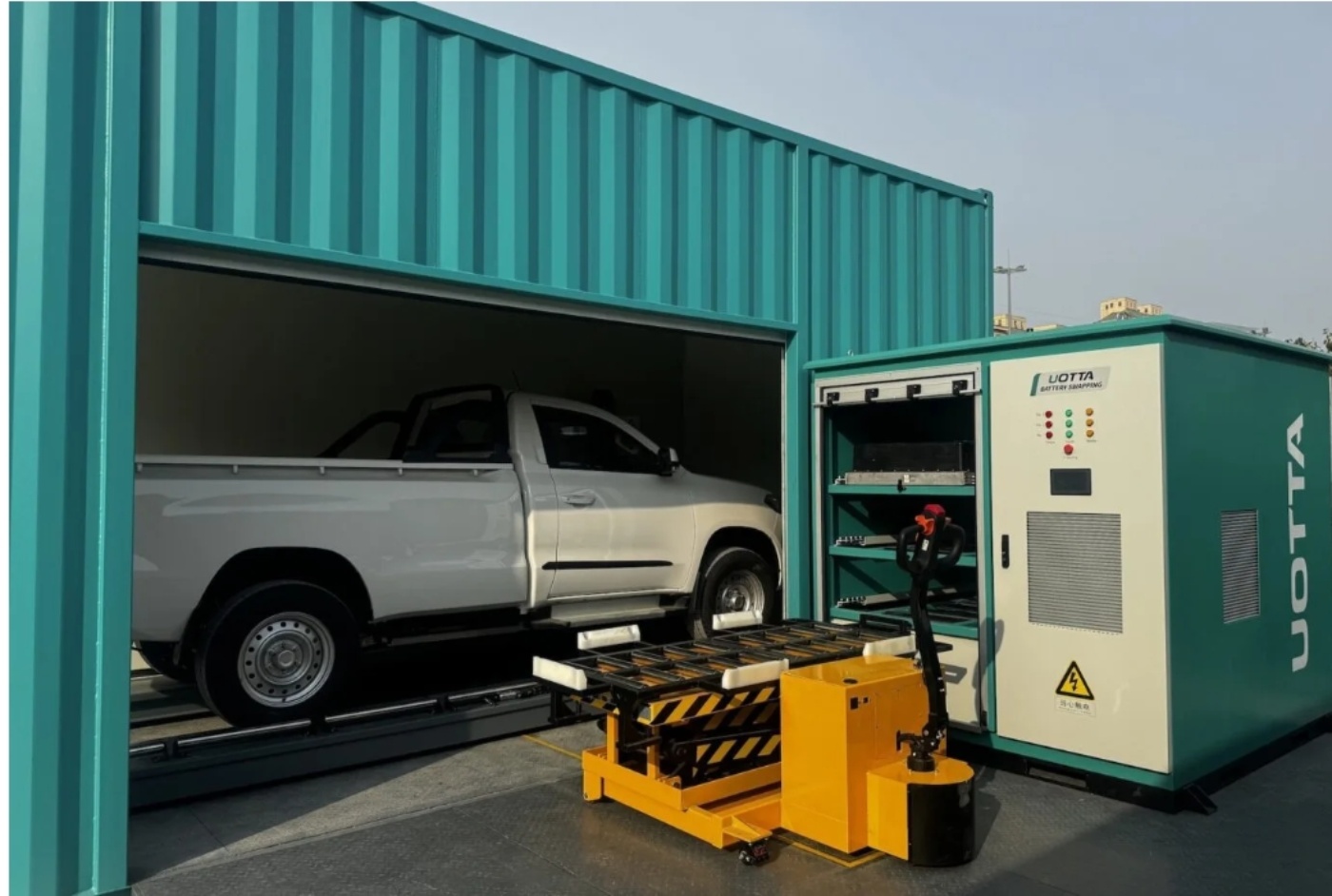A white pickup truck is parked inside a teal-colored container station, next to a UOTTA battery swapping system with a yellow loading platform in front.