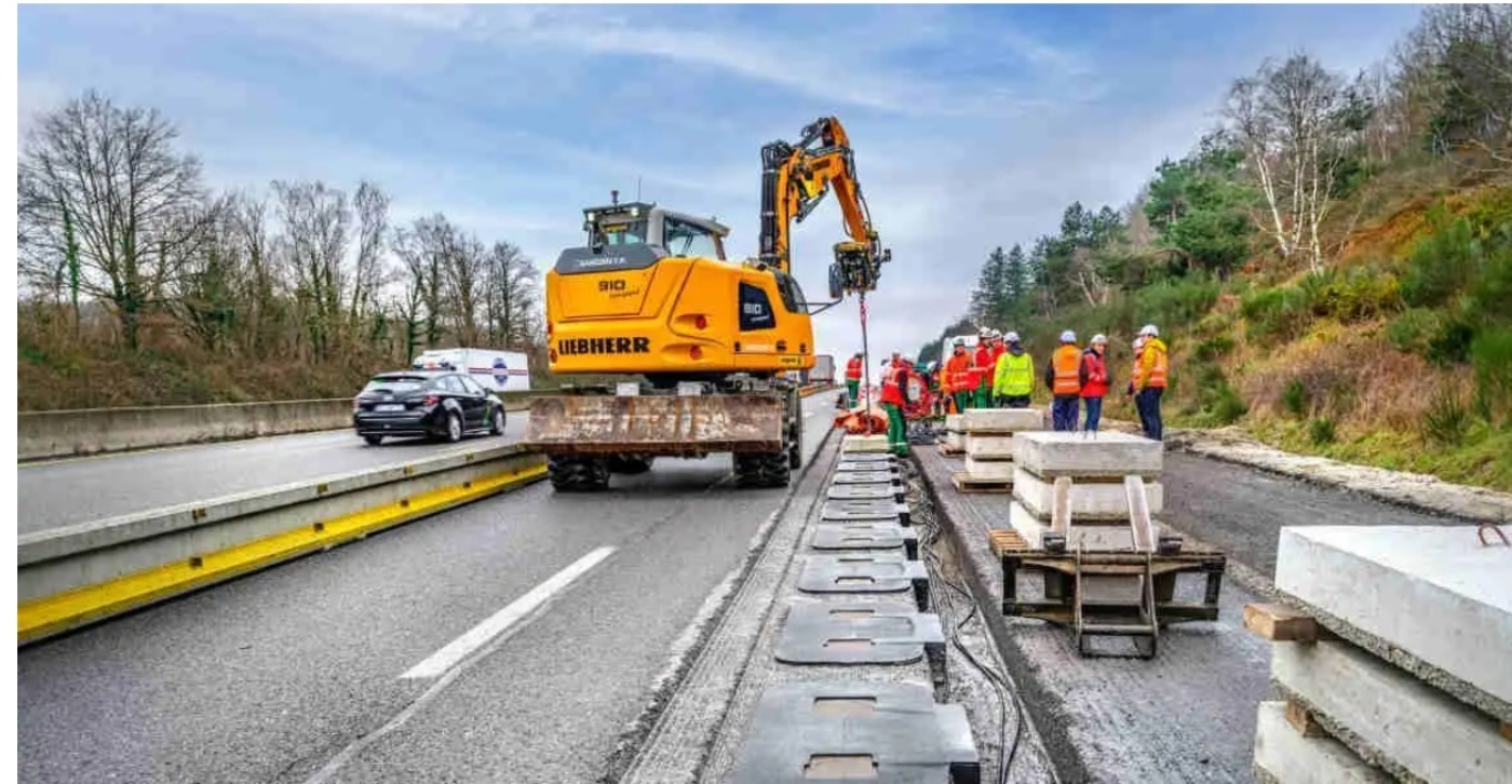 Baustelle auf einer Autobahn mit einem gelben Liebherr-Bagger, der Betonelemente verlegt, umgeben von Arbeitern in Sicherheitskleidung.