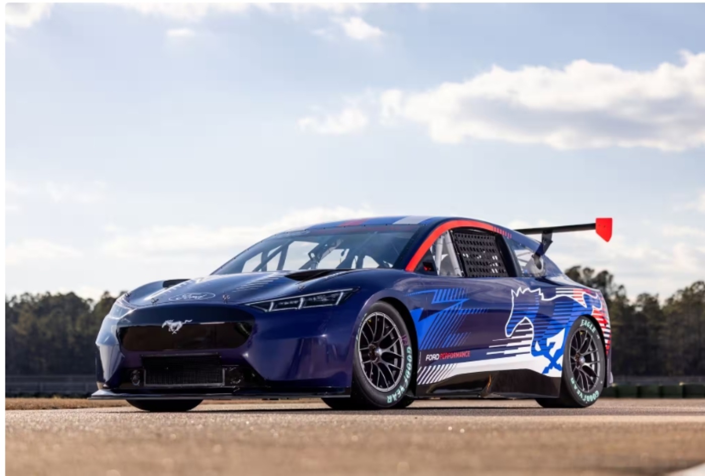 Seitenansicht eines blauen elektrischen Ford Mustang Rennwagens mit Heckspoiler, aufgenommen auf einer Rennstrecke bei klarem Himmel.