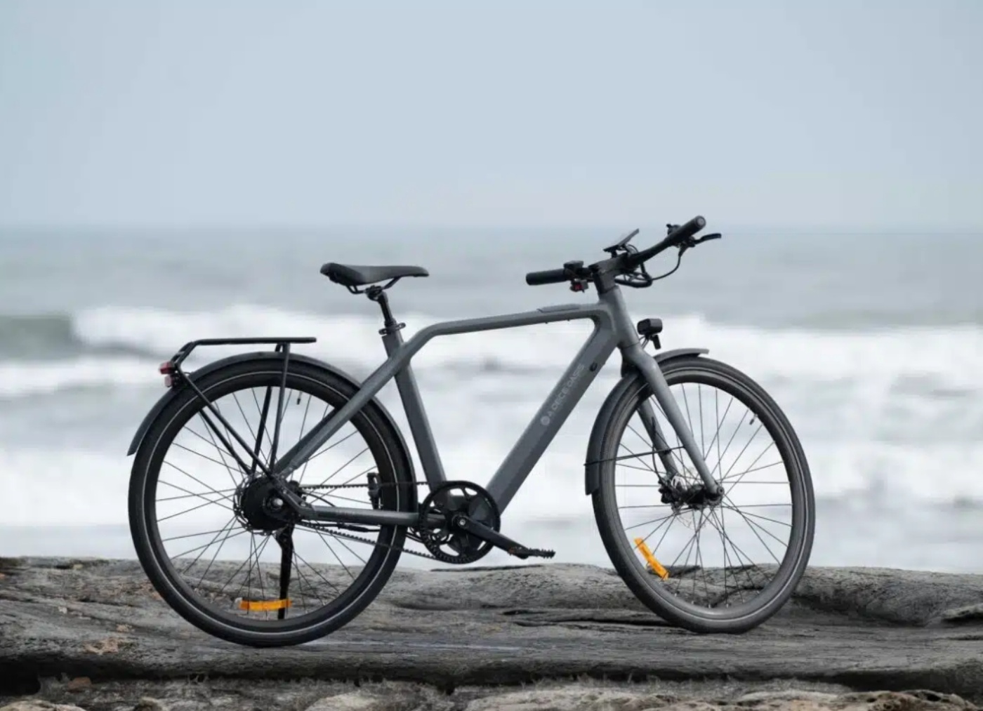Seitenansicht eines mattgrauen E-Bikes von ADO, geparkt auf felsiger Küste mit Wellen im Hintergrund.