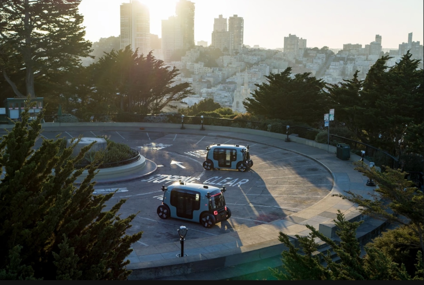Zwei autonome Elektro-Shuttles von Zoox stehen auf einer runden Aussichtsplattform oberhalb einer Stadt bei Sonnenuntergang.