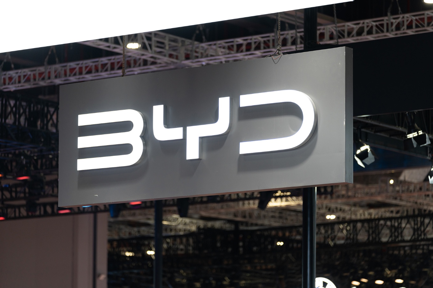 Illuminated BYD logo on a hanging sign at an indoor exhibition booth with lighting trusses in the background.