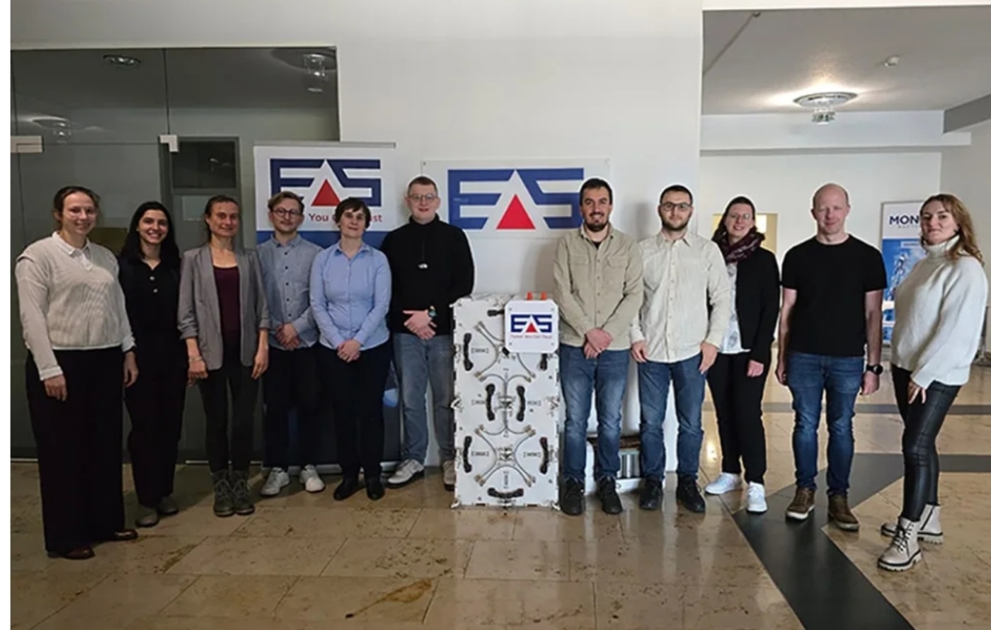 Group photo of eleven people standing indoors around a large white EAS battery module with company logos in the background.