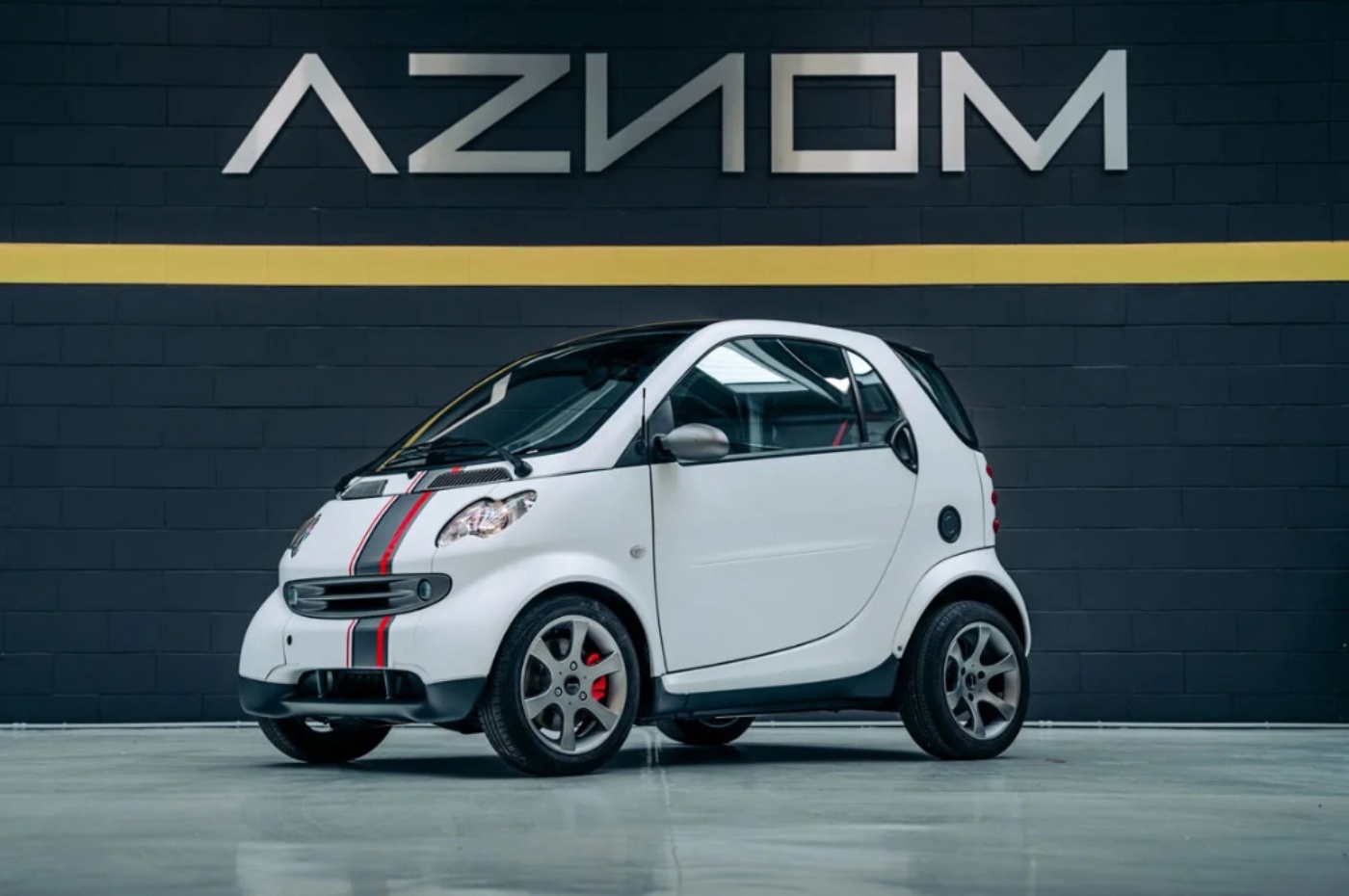 Compact white city car with red and black racing stripes, parked indoors in front of a dark wall with the logo "AZNOM".