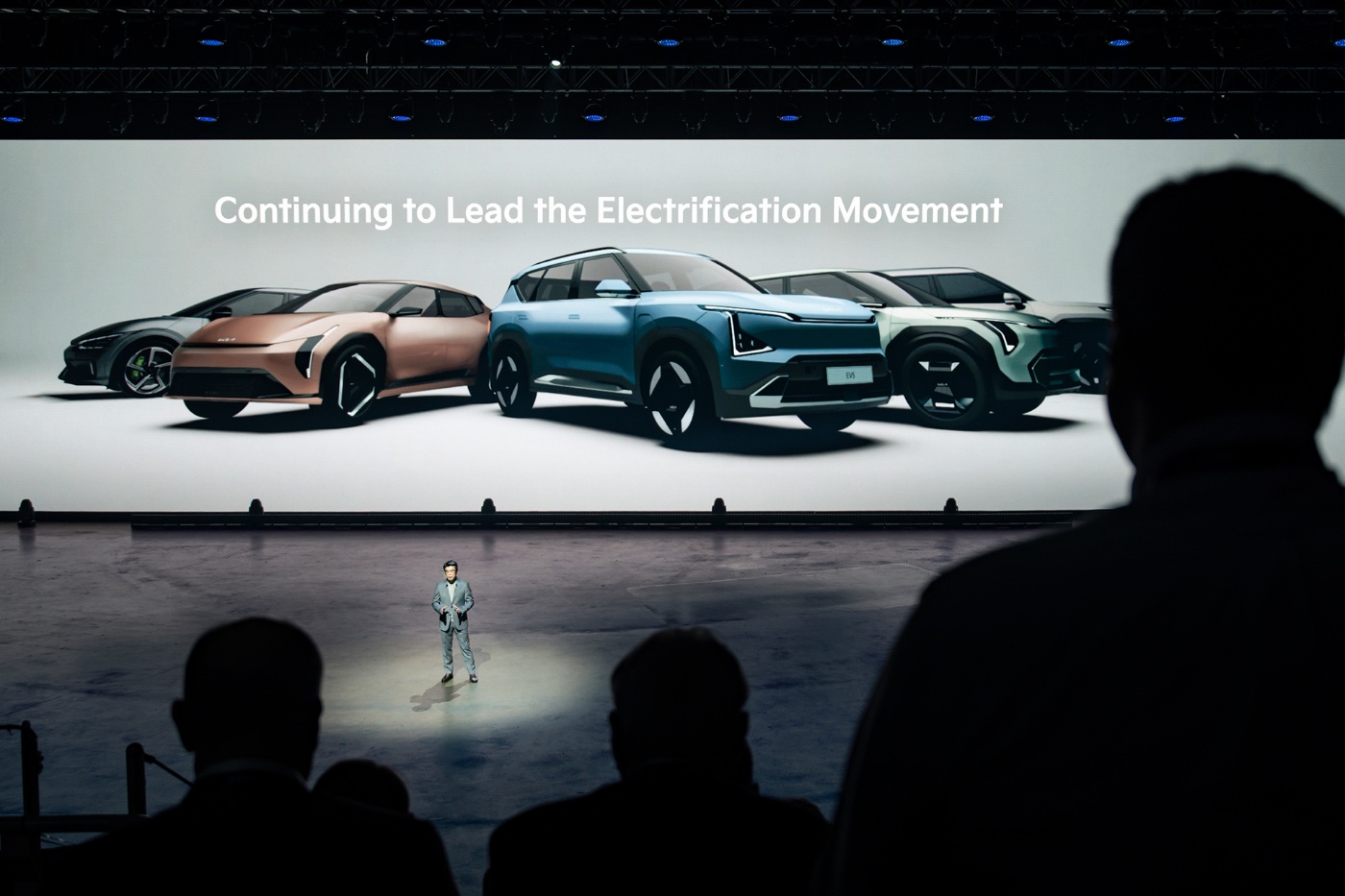 A presenter stands on stage in front of a large screen displaying several Kia electric vehicle models and the slogan "Continuing to Lead the Electrification Movement."