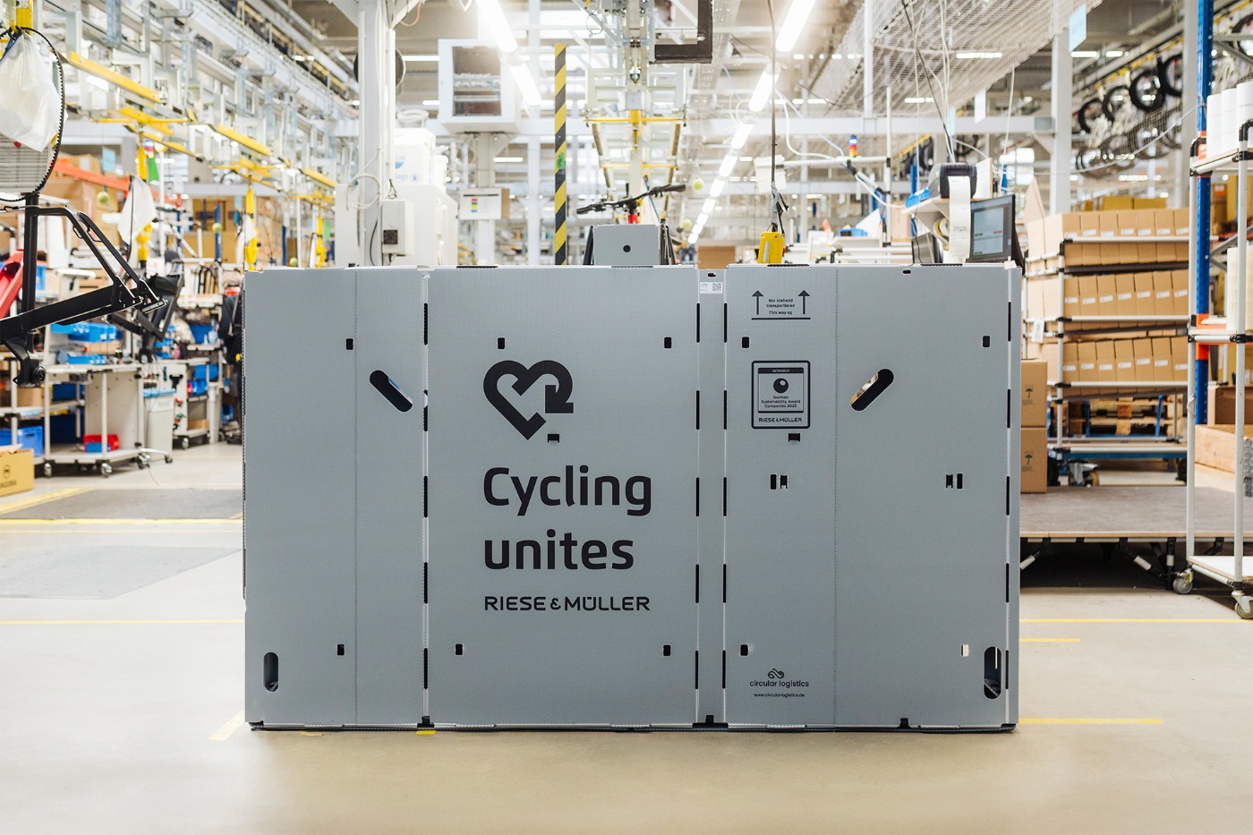 Large reusable shipping box with the slogan "Cycling unites" by Riese & Müller, placed in the middle of a bicycle production facility.