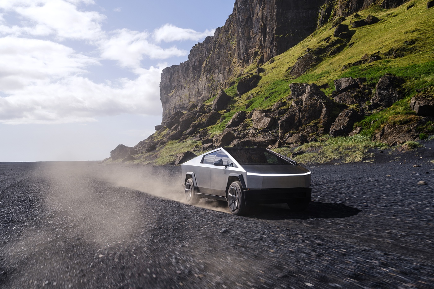 A Tesla Cybertruck drives on a gravel road through a rugged landscape with cliffs and greenery.