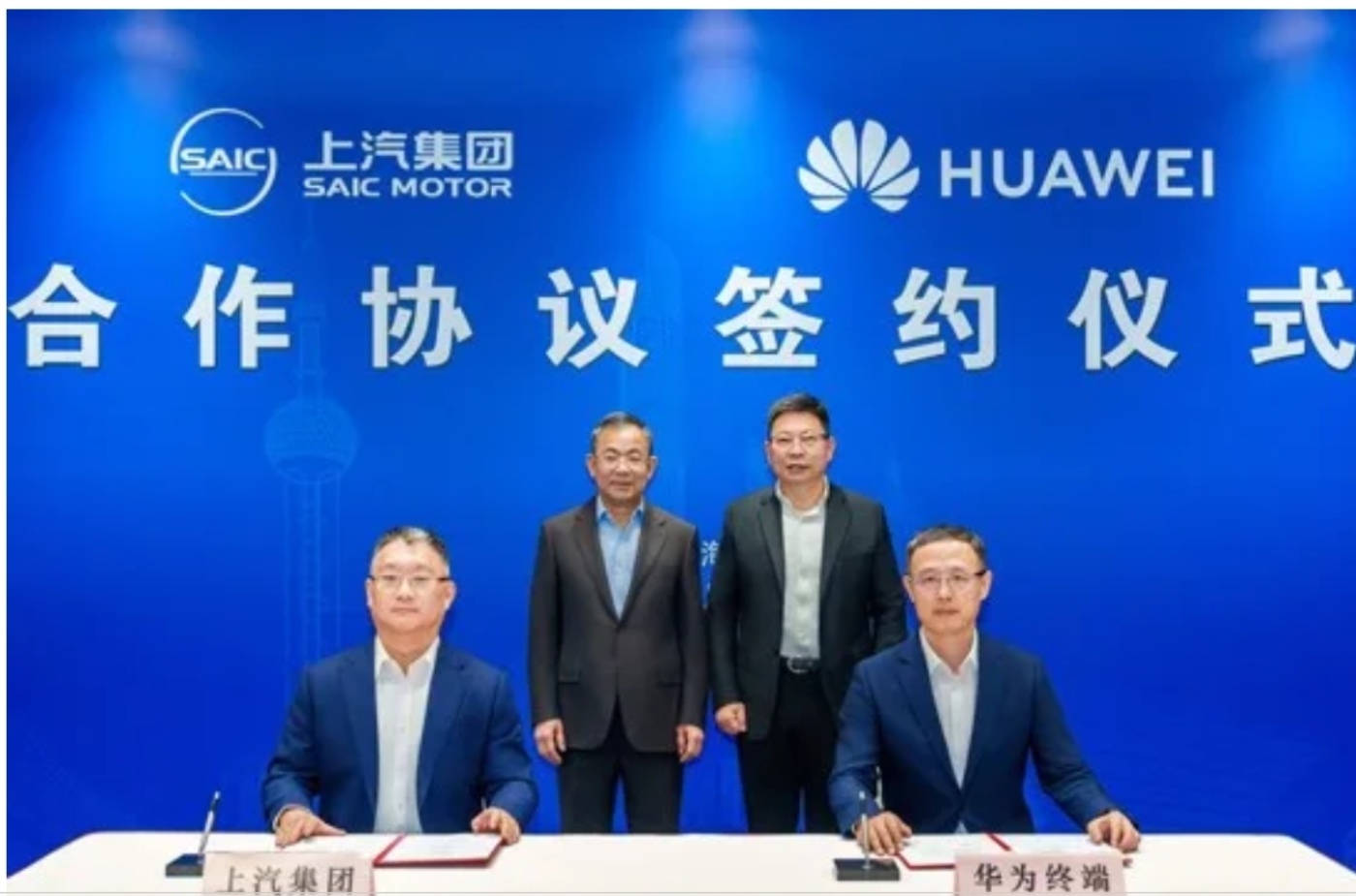 Four men in business attire at a signing ceremony between SAIC Motor and Huawei, with two seated at a table and two standing behind them in front of a blue backdrop.